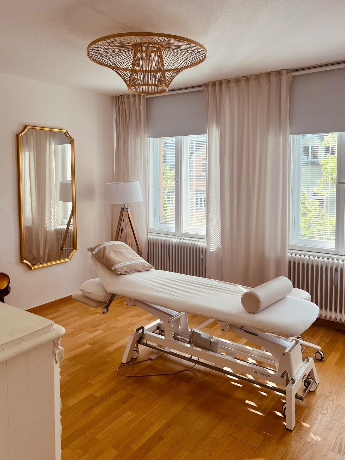 Heller Behandlungsraum der Praxis mit Therapieliege, Spiegel und Rattanlampe
